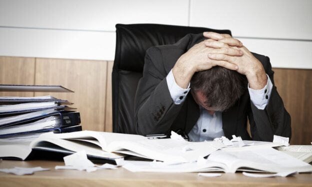 Photo of a worried businessman in dark suit sitting at office desk full with books and papers being overloaded with work, concept of executive drug treatment